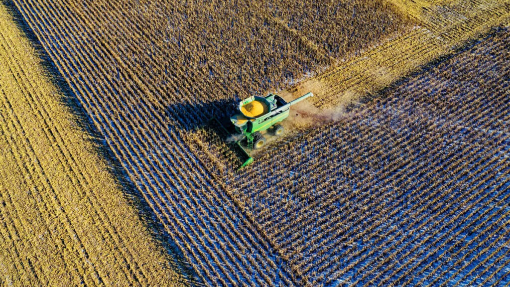 Aerial view of a green combine harvester working in a harvested field, creating parallel rows in the golden and brown crops, akin to the systematic precision brought by Microsoft Cloud Transformation for Insurance technologies.